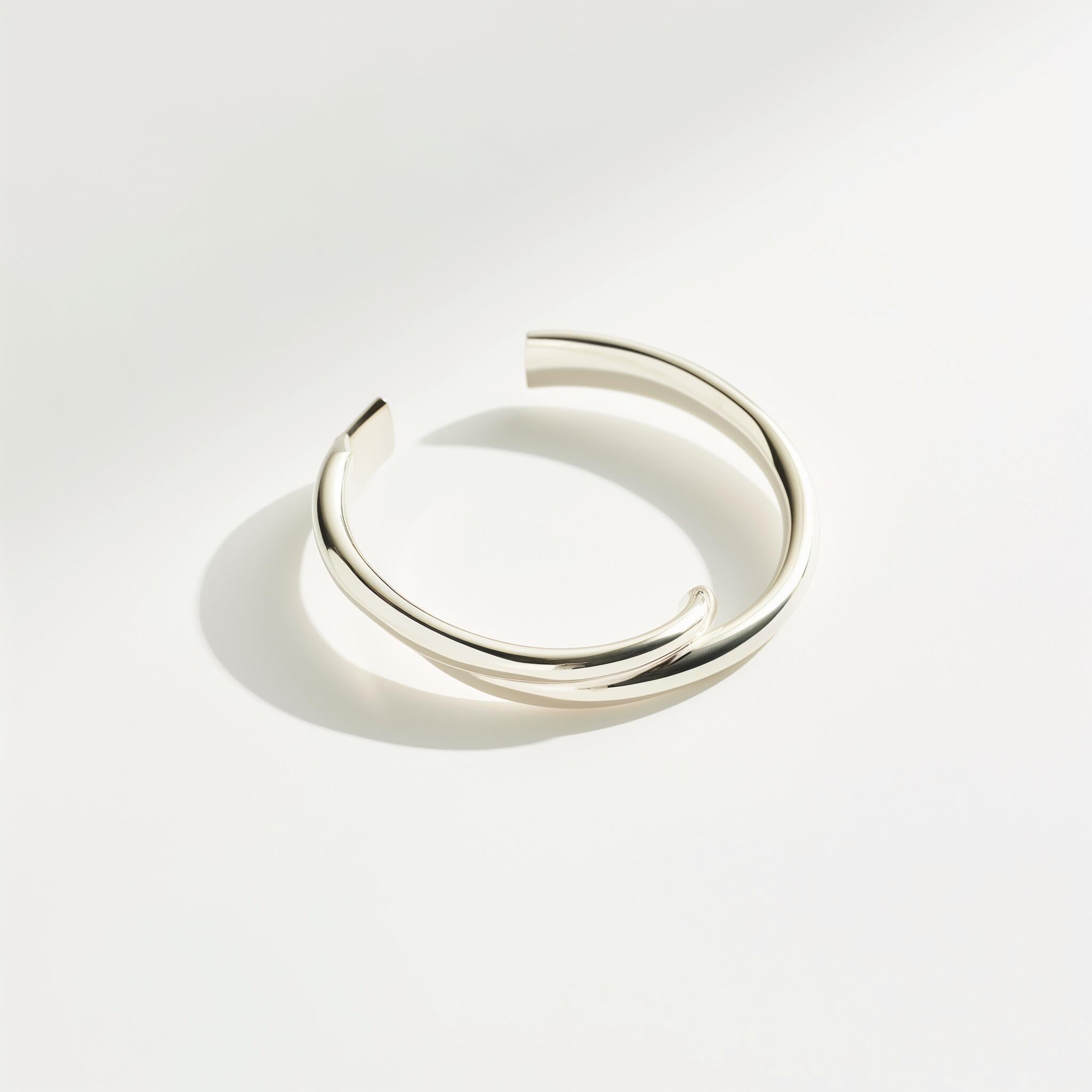 Silver open cuff bracelet on a white background, casting a soft shadow. The minimalist design and smooth finish convey a sense of elegance and simplicity.